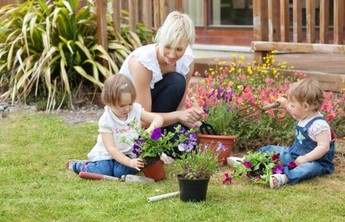 A woman with short blonde hair, dressed in a white top and dark trousers, is kneeling on a well-maintained grassy lawn in a backyard garden. She appears to be engaging with two young children, both with light-colored hair, who are sitting on the grass and planting or tending to potted and flower beds filled with vibrant purple, pink, and yellow flowers. The garden features a neat lawn with a variety of flowering plants arranged close to a wooden deck that has a set of steps leading to the garden area. Behind the group, there are dense ornamental grasses or shrubs, providing a lush green backdrop, and further back, a portion of a house with large windows or sliding doors can be seen, indicating a residential environment in Camberwell. This outdoor space is set in bright, clear weather, with natural sunlight illuminating the scene, emphasizing the healthy grass and colorful blooms, and showcasing a family-friendly gardening space maintained by Gardening Services Camberwell, suitable for outdoor activities and familiar with local garden landscapes in the area.